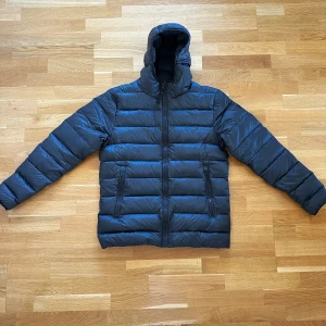 Svart pufferjacka från Helly Hansen - Säljer en svart pufferjacka från Helly Hansen med huva och dragkedja. Jackan har en quiltad design och två fickor framtill. Perfekt för kyliga dagar när du vill hålla dig varm och bekväm.