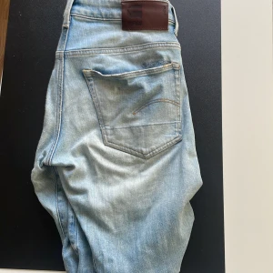 Ljusblå jeansbyxor från G-Star RAW - Säljer ett par ljusblå jeans från G-Star RAW med klassisk femficksdesign och en liten sliten detalj på ena benet. Jeansen har normal passform och raka ben. Perfekta för dig som gillar en avslappnad stil.L32 W32