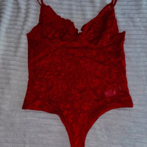 Röd spetsig bodysuit från Bubbleroom - Säljer en röd bodysuit från Bubbleroom i storlek 40. Bodyn är i spets med blommigt mönster, smala axelband och v-ringning. Perfekt för att lyfta vilken outfit som helst med en touch av färg och elegans.