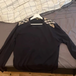 Mörkblå sweatshirt från Burberry med rutiga axlar - Säljer en mörkblå sweatshirt från Burberry med klassiska beige, vita och svarta rutiga detaljer på axlarna. Tröjan är i 100% bomull och har en avslappnad passform med långa ärmar. Perfekt för en stilren och bekväm look.