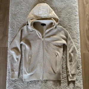 Beige cardigan med luva - Säljer en beige huvjacka med hel dragkedja framtill och två fickor med dragkedja. Jackan har en quiltad huva och är tillverkad i ett strukturerat material. Perfekt för lager-på-lager och snygg till vardags. Från Indicode, storlek Small.