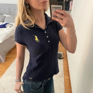 Mörkblå pikétröja från Ralph Lauren - Säljer en klassisk mörkblå pikétröja från Ralph Lauren med gul broderad logga på bröstet. Tröjan har korta ärmar, krage och knappar framtill. Tröjan är i barnstorlek men passar mig som oftast bär XS och är 165cm