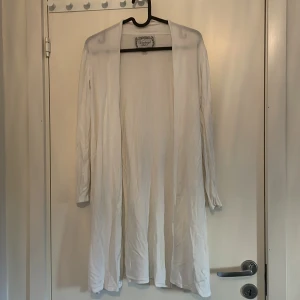 Lång kofta - Vit lång kofta. St 146/152. Har två hål vid varsin arm  som är insydda men syns inte så mycket. Säljer för att den tyvärr inte passar längre. Har en sluts vid sidan. Väldigt fin på! 🤍👚
