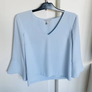 V-ringad blus från H&M storlek 36 - Märke: H&M Vad: En v-ringad blus i vävd kvalitet. Blusen har trekvartslång trumpetärm med slits. Storlek: 36 Upplevd i storlek: Normal Färg: Ljusblå Material: 100% polyester  Skick: Inget att anmärka på