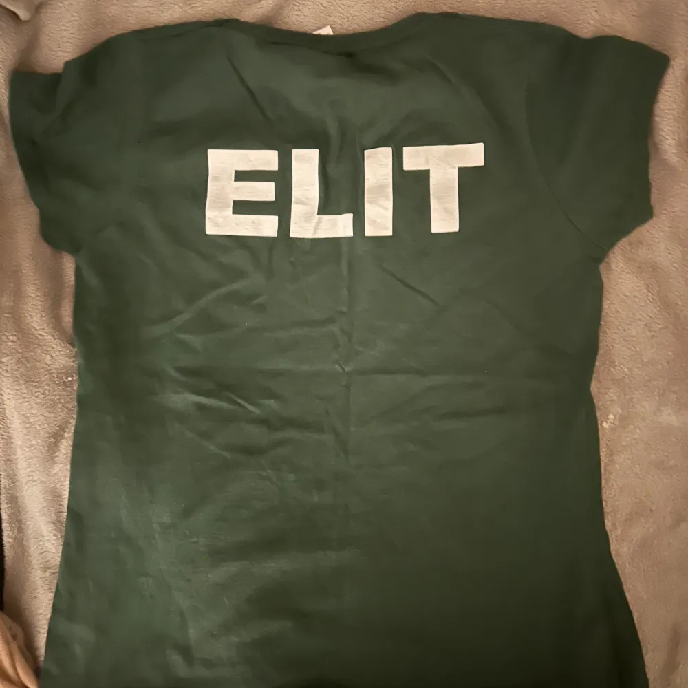 En skogsgrön t-shirt där det står ”elite” på baksidan, samt en ribbad beige t-shirt med volang vid midjan samt axlarna. 20 kr st, 30 kr tsm.. T-shirts.