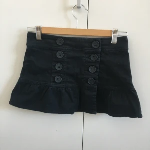 Svart minikjol - Min syrras svarta minikjol i jättefint skick. Det är en omlottkjol som går att öppna helt och knäpps med knapparna. Liten i storleken, gissar XS eller XXS! Längd: 27,5 cm, midja: 33,5 cm. Skriv för fler bilder!