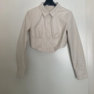 Croppad skjorta i läderimitation- ny! - Superfin skjorta i läderimitation i en beige/vit färg! Lappen finns kvar och har endast varit provat. Har en väldigt liten fläck på insidan (som inte syns när den är på), kan skicka fler kort på önskemål ❤️