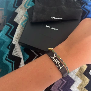Ysl armband  - Säljer mitt fina ysl armband i svart med silver detaljer. Box och dustbag medföljer köpt på NK för 2800kr. Knappt använd. Storlek M (16 cm) 
