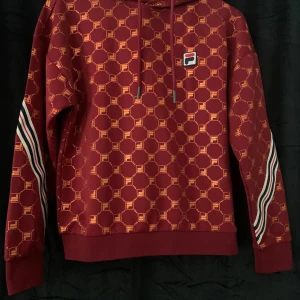 Exklusive huvtröja hoodie FILA vinröd med guldmönster strl M - Exklusive huvtröja hoodie FILA vinröd med guldmönster strl M Superfint skick. Yttertyg: 94% Polyester, 6% Elastane Foder: 100% Polyester