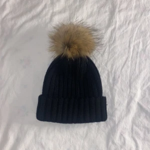 Svart mössa med pälsboll (äkta)  - Säljer en jättefin svart mössa med en pälsboll där pälsen är äkta. Mössan är knappt använd och i nyskick. 