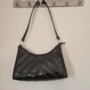 Handväska - Svart och glansig handväska med mönster. Guldfärgad dragkedja. Bara använd ett fåtal gånger. I mycket bra skick.