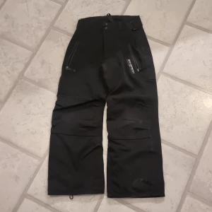 Termobyxa/skidbyxa - Svart termobyxa/skidbyxa med fickor som har dragkedja. Hällor i midjan och kan justeras med kardborreband. Liten "krok" på en benet nedtill som saknas. 