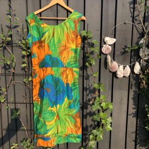 Färgstark 60-talsklänning!  - Hemsydd. 60-tal. Snyggt skärp till med knytbar rosett. Slitsar i sidorna. Fodrad i kjoldelen. Bystinsnitt. Dragkedja bak. Byst: 48x2 Midja: 39x2 Stuss: 45x2 Längd: 105 cm  Ärmhål: 22x2