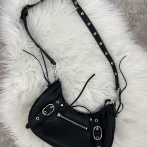 Svart handväska - Svart handväska från H&M med silvriga detaljer. Man kan justera bandet på vöskan. Dragkedja och mycket utrymme. Knappt använd.