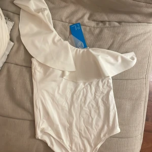 Ny baddräkt - Helt oanvänd vit one-shoulder baddräkt med volang. Prislappen finns kvar. Originell pris 29,99€