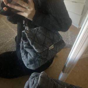 Grå denim väska - Fin grå denim väska med 2 uppdelningar inuti samt dragkedje ficka. Man kan välja att ha den runt sig på axeln eller under armen. Köpt för 390kr.