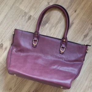 Vinröd stor väska  - Helt ny. Perfelt till skola eller jobb då den är rymlig, har massa fack och dragkedja. Korta band plus ett långt band som går att sätta på eller ta av. Ett litet märke på botten som knappt syns.