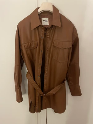 Skjorta i skinnimitation  - Skjorta i skinnimitation från Zara. Den kommer med ett bälte som man spänner runt midjan. Endast använd 3 gånger och är i mycket bra skick. Upplever att den är normal i storlek. Kan bäras som en skjorta eller som en jacka när det är lite varmare ute. 