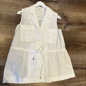 Blus - Blusen har krage och slag samt dold knäppning fram.  Bröstflickor med lock och dragsko i midjan. Aldrig använd. 