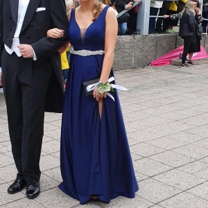 Balklänning - Balklänning som är använd endast en gång, passar person som är 165 + ca 8cm klackar. Klänningen är marinblå med ett bälte av stenar, v-ringning både fram och bak. Slits på vänster sida.