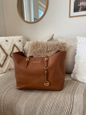 Äkta Michael Kors väska - Äkta Michael Kors Jet set Medium Saffiano Leather Tote Bag i färgen cognac/brun.  I använt men fint skick.  Endast lite sliten på handtagen (syns på bilden) och går lätt att rusta upp hos en skomakare om man vill det.   Rymmer allt och lite till.