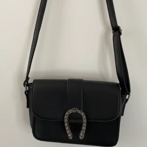 Väska - Svart väska med silver detalj på framsidan, lagom liten med 2 fack inne (ett stort och ett mindre med dragkedja) och med axelband.