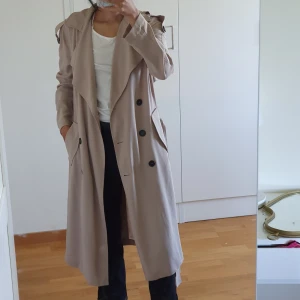 Beige kappa - Gina tricot. Bra skick förutom att översta knappen saknas och att snöret vid ena sidan har gått av men går att sy ihop. Plaggets längd 113 cm och ärmlängd 58 cm.