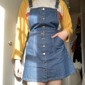 Hängselklänning Jeans - Hängselklänning i jeanstyg med knappar längs hela framsidan, två stora fickor fram och justerbara axelband.