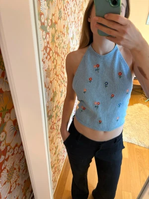 Blå stickad halterneck topp med blommor - Ljusblå stickad halterneck topp med broderade små blommor i olika färger. Toppen är ärmlös, croppad och knyts i nacken och ryggen. 