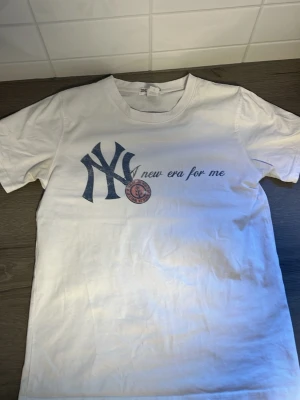 Vit t-shirt från 2K WRLD med tryck - Vit t-shirt från 2K WRLD med stort tryck framtill som visar NY-logga och texten 'new era for me'. Tillverkad i 100% bomull och har klassisk rund hals och korta ärmar. Perfekt för dig som gillar streetwear och enkel stil.