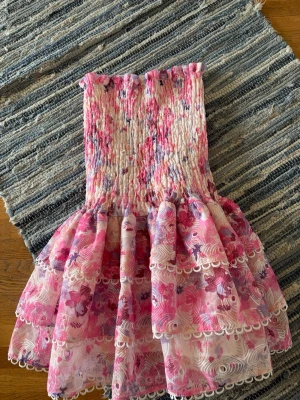 Blommig kort klänning Designby Amor - Supersöt kort klänning från Designby Amor i blommigt mönster med rosa, lila och vita toner. Klänningen är axelbandslös med smockad överdel och volangig kjol med spetsdetaljer. Perfekt för sommarens alla tillfällen!