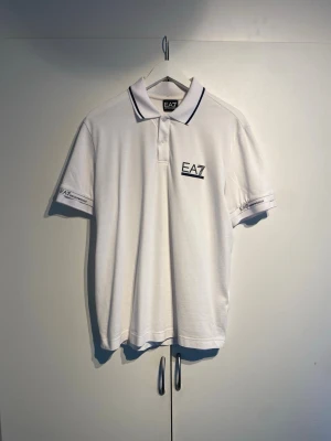  EA7 Emporio Armani Pike - Klassisk vit pikétröja från EA7 Emporio Armani med svart logga på bröstet och ärmarna. Tröjan har krage med tunna svarta ränder och knäppning framtill. Tillverkad i mjuk bomull för en bekväm känsla. Knappt använd, väldigt bra skick. Nypris 1800kr.
