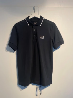  EA7 Emporio Armani pike  - Klassisk svart pikétröja från EA7 Emporio Armani med vit logga på bröstet och vita detaljer på kragen. Tröjan har korta ärmar, knappar framtill och är tillverkad i mjuk bomull. Perfekt för en clean och stilren look. Väldigt bra skick, knappt använd. 