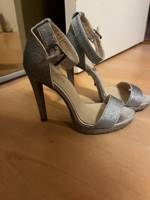 Glittriga silverfärgade pumps - Säljer ett par glittriga silverfärgade pumps med hög klack och öppen tå. Skorna har en bred rem över tårna och justerbart ankelband med spänne. Perfekta för att lyfta vilken outfit som helst med extra bling.