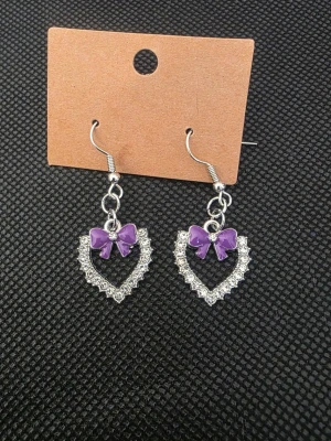Hjärformade örhängen med lila rosett - Säljer ett par söta örhängen i form av hjärtan dekorerade med glittrande stenar och en lila rosett upptill. Örhängena har silvrig metall och hängande krok. Perfekta för dig som gillar gulliga accessoarer med lite extra bling.