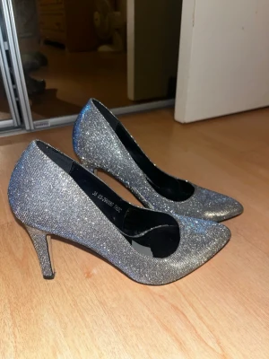 Glittriga silverfärgade pumps - Säljer ett par glittriga pumps från VOX Shoes i silver med spetsig tå och smal klack. Skorna har en elegant form och är täckta av skimrande material som fångar ljuset. Perfekta för att sticka ut på festen eller till en snygg outfit. Materialet på sulan är känsligt  och går av om det blir friktion. så säljer de mycket billigare. 