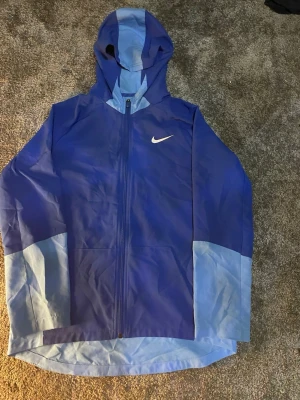 Blå Nike vindjacka med huva - Snygg blå vindjacka från Nike med dragkedja framtill och huva. Jackan har ljusblå detaljer på ärmar och huva samt en vit Nike-logga på bröstet. Perfekt för blåsiga dagar och sportiga outfits.