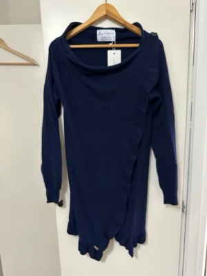 Mörkblå omlottklänning Evy Alfrida - Mörkblå långärmad klänning från Evy Alfrida med omlottdesign och asymmetrisk nederkant. Klänningen har en bred krage och är tillverkad i ett mjukt material som ger en snygg siluett. Perfekt för dig som gillar unika detaljer och svensk design.