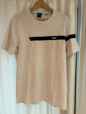 Vit t-shirt från BOSS med tryck - Snygg Vit t-shirt från BOSS med svart drag och logga över bröstet. Klassisk rund halsringning och korta ärmar. Perfekt till jeans eller shorts för en clean och stilren look.