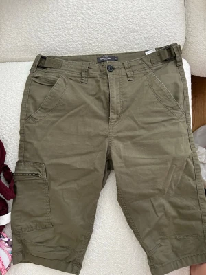 Olivgröna cargoshorts med fickor - Säljer ett par olivgröna cargoshorts med flera praktiska fickor, bland annat sidoficka med dragkedja och bakfickor med lock. Klassisk passform och bälteshällor. Perfekta för sommaren och avslappnade dagar.