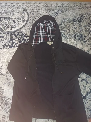 Svart hoodie från Burberry med rutigt foder - Säljer en svart hoodie från Burberry med dragkedja och klassiskt rutigt foder i huvan. Hoodien har dragsko i huvan och diskret Burberry-logga broderad på bröstet. Perfekt för dig som gillar stilrena och exklusiva plagg.