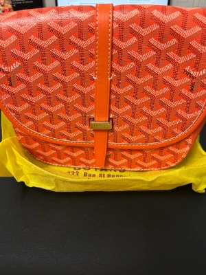 Orange axelväska - Säljer en orange axelväska med det klassiska mönstret och silverfärgad spänndetalj. Väskan har en rundad form och är tillverkad i canvas med läderdetaljer. Perfekt för dig som vill sticka ut med en färgstark accessoar.