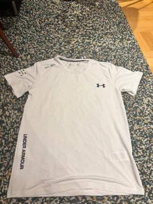 Vit tränings-tshirt från Under Armour - Snygg vit tränings-tshirt från Under Armour med svart logga på bröstet och text längs sidan. Tillverkad i lätt och snabbtorkande material, perfekt för löpning eller gym. Rund halsringning och korta ärmar för maximal rörelsefrihet.