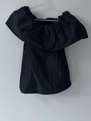 Svart offshoulder topp från COS - Säljer en svart offshoulder topp från COS i storlek 34. Toppen har en bred volang runt axlarna som ger en statement-look och är ärmlös. Perfekt för dig som vill ha en stilren men ändå unik topp.
