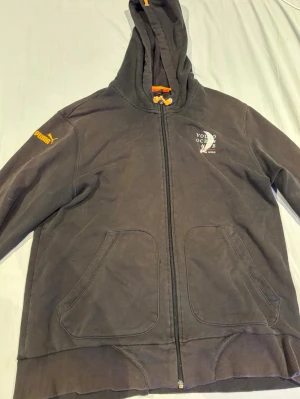 Svart zip-hoodie från Puma - Säljer en svart hoodie från Puma med dragkedja, huva med orangea snören och två stora fickor framtill. Puma-logga på ärmen och tryck på bröstet. Perfekt för en avslappnad och sportig stil.