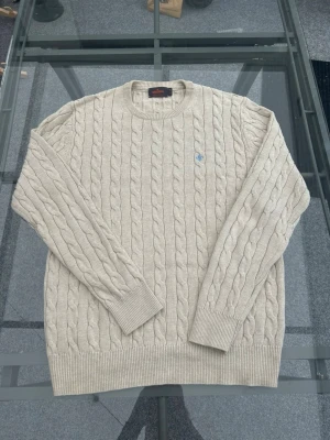 Morris Stockholm Kabelstickad Tröja – Beige Pima Cotton (Storlek M) - Säljer en klassisk och stilsäker kabelstickad tröja från Morris Stockholm i en fin ljusbeige/off-white färg. Den är tillverkad i 100% ljuvlig pima-bomull, vilket gör den otroligt mjuk, skön och av högsta kvalitet. Tröjan har en rund halsringning och pryds av Morris klassiska broderade lilja på bröstet i en snygg ljusblå kontrast. Ett perfekt preppy-plagg som är lika snyggt över en skjorta som för sig själv.  Märke: Morris Stockholm  Modell: Kabelstickad tröja / Cable Knit  Storlek: M (Herr)  