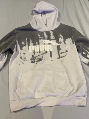 Gråvit hoodie med tryck från Puma - Säljer en gråvit hoodie från Puma med coolt svartvitt mönster och stor logga på bröstet. Hoodien har huva och en stor magficka. Perfekt för dig som gillar streetwear och vill sticka ut lite extra.