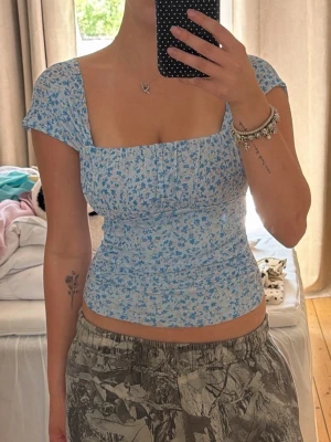 Blommig blå croppad topp - Söt croppad topp med blått blommönster och korta ärmar. Toppen har en fyrkantig ringning och sitter tight mot kroppen. Perfekt för dig som gillar romantisk och trendig stil.