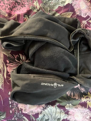 Svart hoodie med dragkedja Sail Racing - Svart hoodie från Sail Racing med dragkedja framtill och huva. Tillverkad i mjukt material med fleecefoder på insidan för extra komfort. Logga på ena ärmen. Perfekt för en avslappnad och sportig stil.