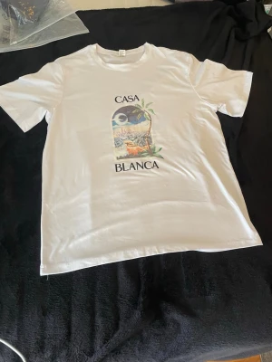 Vit t-shirt med Casa Blanca-tryck - Säljer en vit t-shirt med färgglatt tryck och texten 'Casa Blanca' på bröstet. T-shirten har rund hals och korta ärmar. Perfekt för dig som gillar stilrena plagg med en twist.